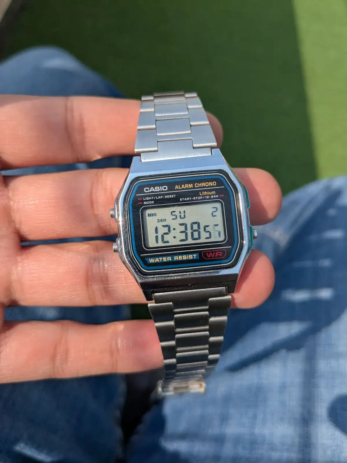 Casio A158W plateado con correa de acero sostenido en la palma de una mano, sobre un vaquero
