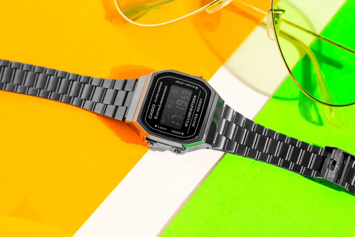 Casio digital con correa de acero sobre un fondo amarillo y verde, junto a unas gafas